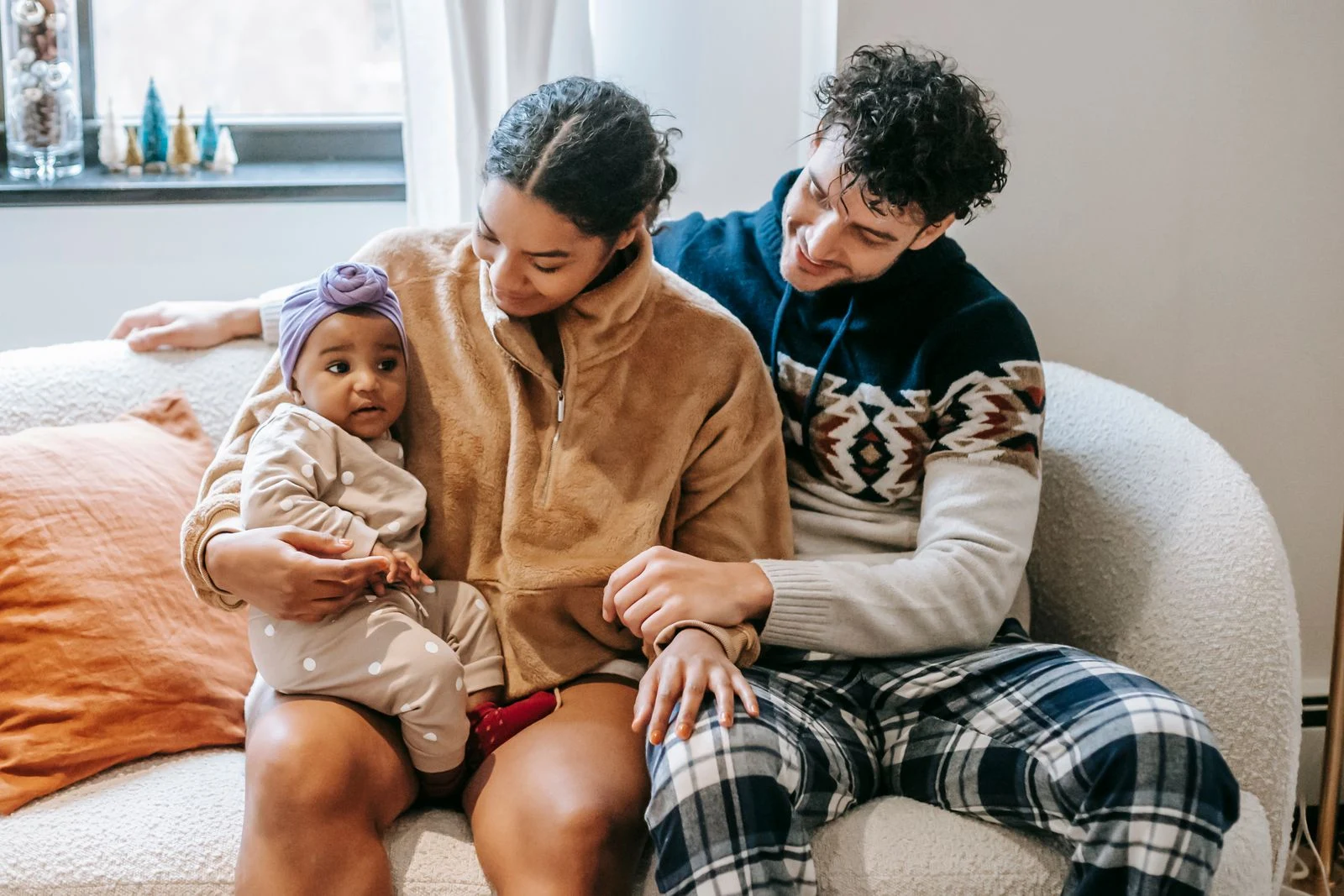 Two adults sitting close together on a sofa, smiling toward a baby held in their arms.